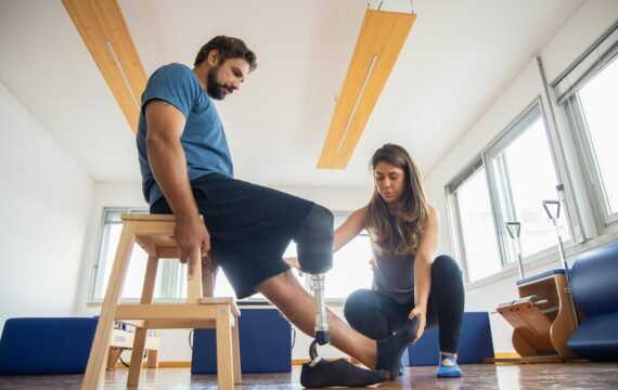A trainer assists a man with a prosthetic leg during indoor physical therapy.